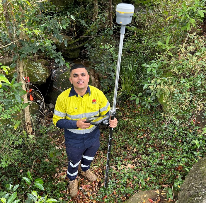 Nick Baran wears a high-visibility yellow and navy work uniform with reflective stripes. He is standing in a lush, wooded area holding surveying equipment.