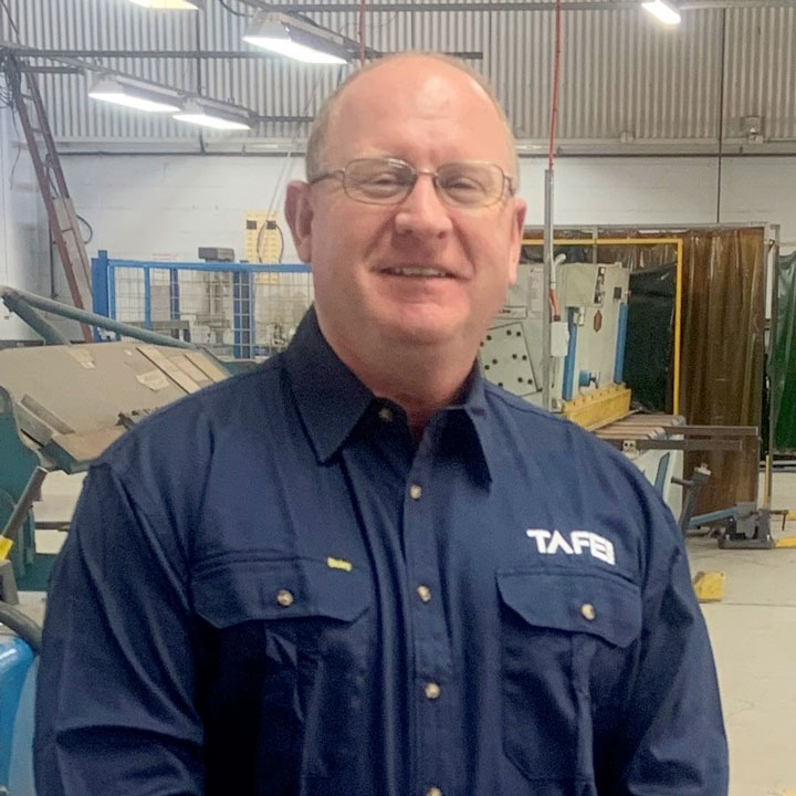 The image shows Stuart McKinder in a dark blue TAFE NSW button-up shirt. He is wearing glasses and smiling at the camera. He is in a warehouse setting.