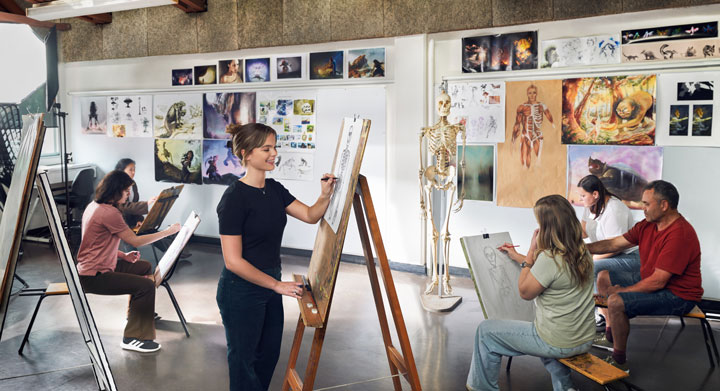 A group of students are in an art workshop sketching a skeleton model that is propped on a stand at the front of the class. There are lots of art works plastered on the walls around them.