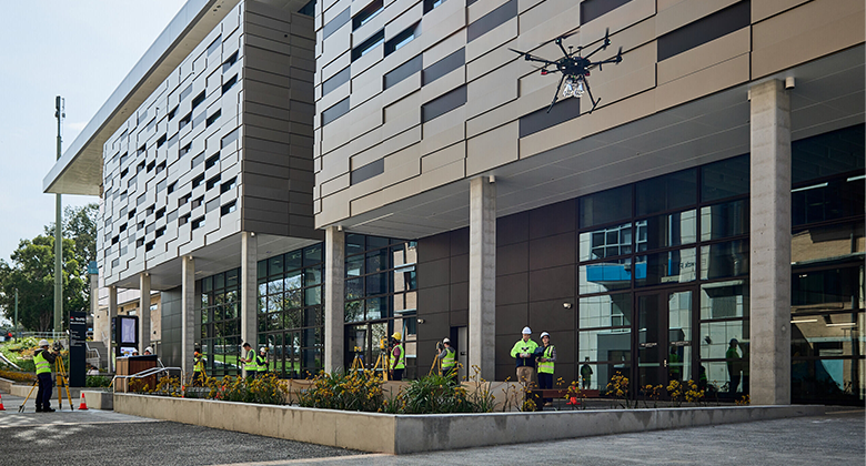 People standing outside a building dressed in high-viz gear, surveying the building. One of the workers is flying a drone in the foreground. 