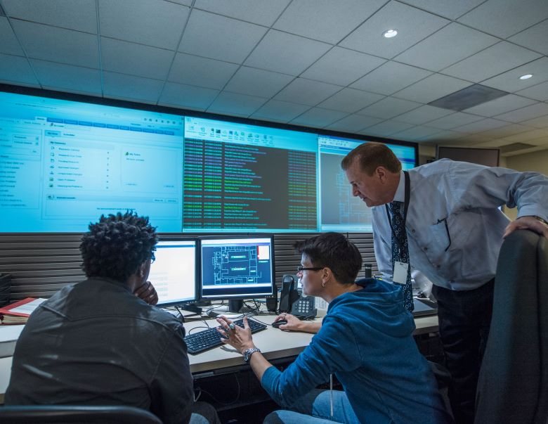 A TAFE NSW cyber defence classroom with a teacher, two students on computers, and a large screen showing course data.