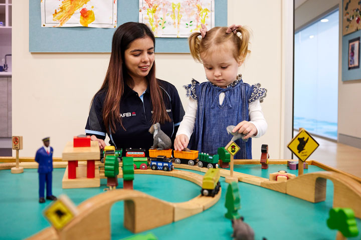 A student studying early education sits with a toddler while the little girl plays with a toy railway set. The little girl wears high pigtails with pink ribbons and appears engaged in the activity.