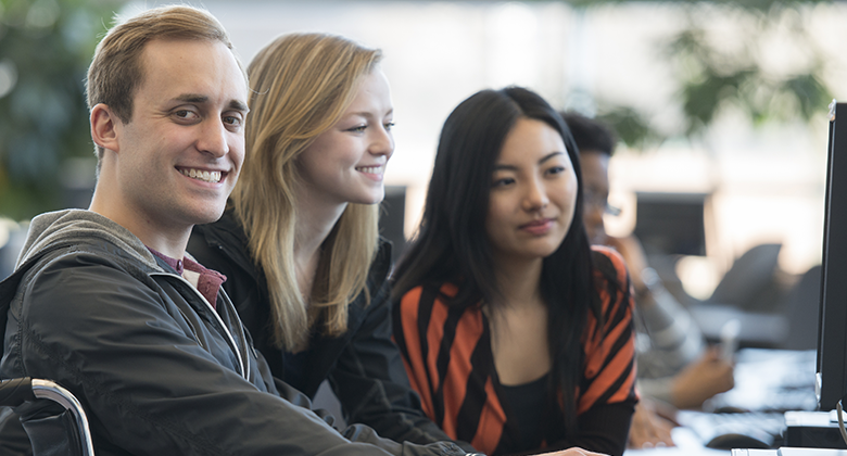 Three students working together at a computer, smiling, with one of them in a wheelchair.
