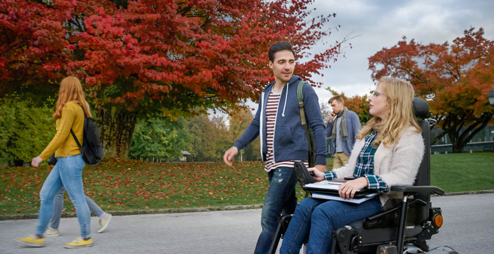 Two students conversing together on a park path. The young man is walking and the young woman is in a motorised wheel chair.