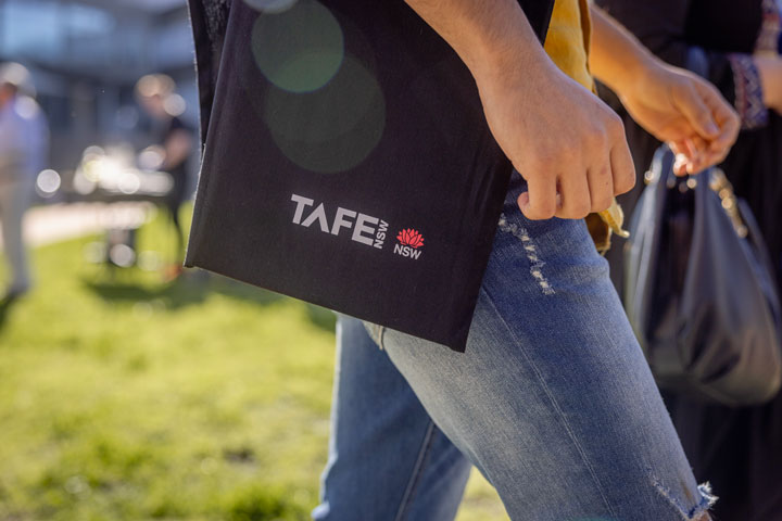 A student is wearing ripped jeans and a black tote bag displaying the TAFE NSW logo. The student is walking outside with another person also carrying a black bag. A blurred TAFE NSW campus and 2 other people are seen in the background. 