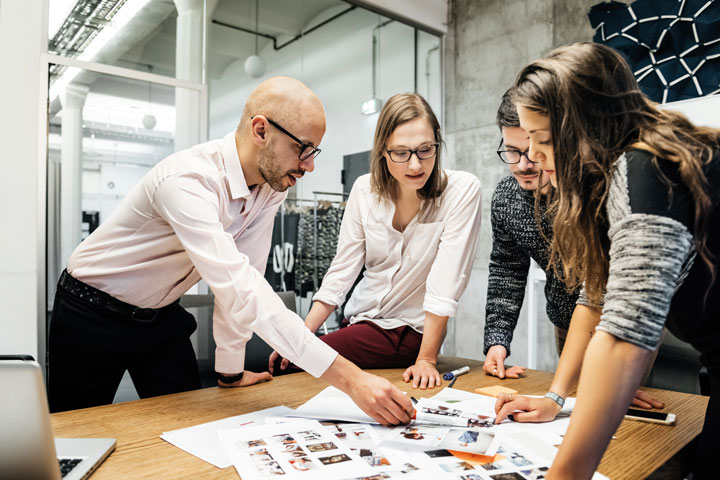 A diverse group of four professionals gather around a table  in a modern office space. The table is covered with documents, fashion images and photos. They are physically engaging in the material while discussing ideas.