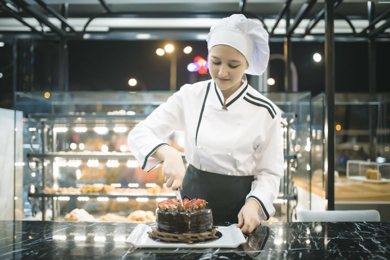 Professional pastry chef slicing a decadent chocolate cake topped with fresh strawberries. She is standing in a modern bakery with illuminated glass displays showcasing a variety of baked goods.