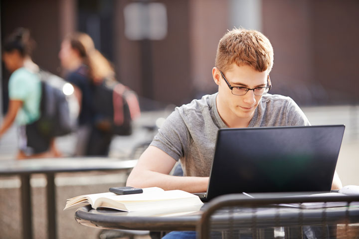 A young man with glasses working on a laptop at an outdoor table, with a smartphone and open book beside him. Blurred figures of other people with backpacks walk by in the background.