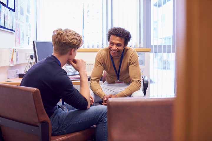 A young man is seated in an office, engaged in a one-on-one conversation with a smiling older man who is a counsellor or advisor offering support and guidance. The older man is wearing a lanyard and is leaning forward, creating a welcoming and attentive posture. The office is well-lit with natural light coming through large windows, and there are computer equipment and documents on the desk. 