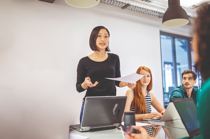 A women with short hair stands at the front of a large table with her seated colleagues. She is presenting an idea or information, whilst holding a piece of paper in one hand and has a laptop screen on the desk in front of her. 