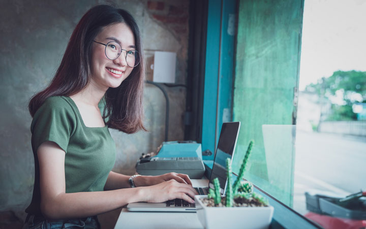 A young woman with round-rimmed glasses looks happy while sitting at her desk using her laptop. Her desk faces a street-facing window. A potted cactus is on the desk in the foreground.