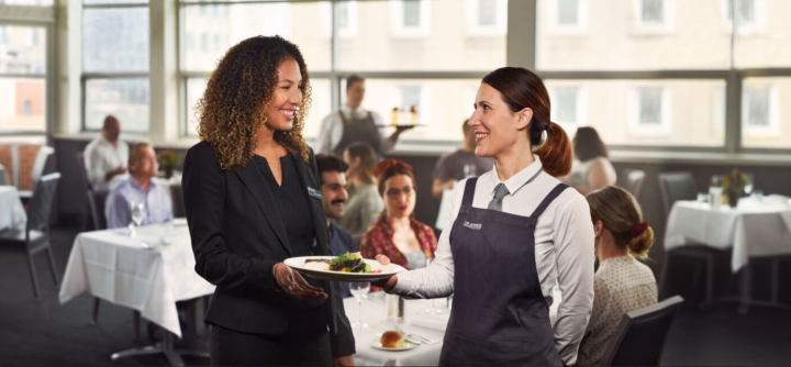 The image shows a professional interaction in a restaurant setting. A smiling server in a uniform, holding a plate of food, is handing it to a woman in business attire. The two appear to be exchanging polite conversation. In the background, diners are seated at tables, engaged in their meals, while other staff members work. 
