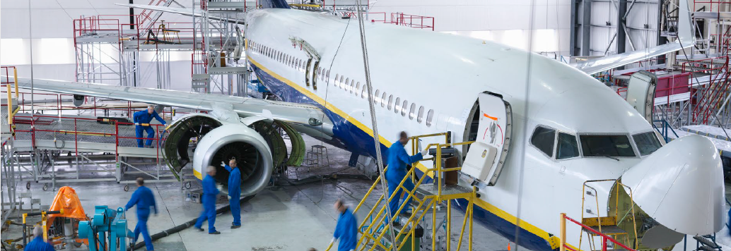 The image shows a large plane in a warehouse that is being services by people in blue uniforms. The workers are slightly blurred, indicating that they are moving around and working on the plane.