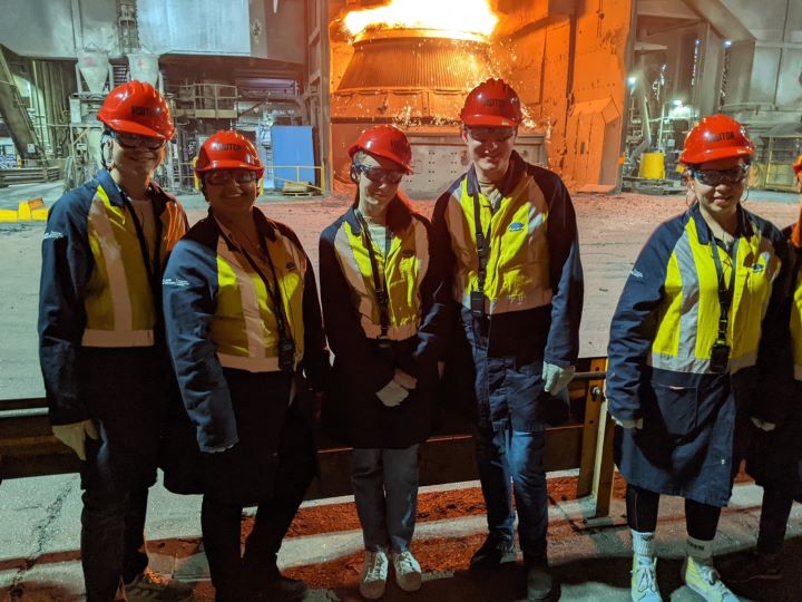 The image shows five people posing for a photo in a steel manufacturing facility. They are wearing protective clothing including long pants, Hi-Vis jackets, Hard Hats and sunglasses. They look happy to be there and to be learning.