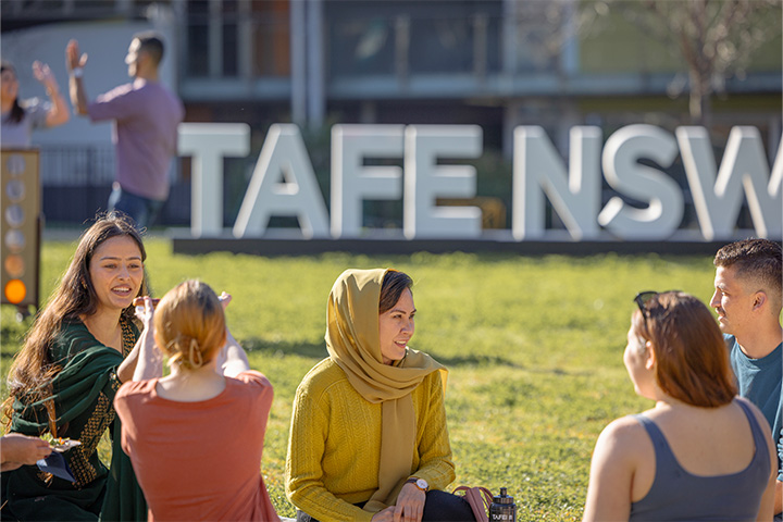 A group of students sitting on the grass and chatting in front of a large "TAFE NSW" sign. The group includes a diverse mix of individuals, with one wearing a hijab. Two people in the background are giving a high-five.