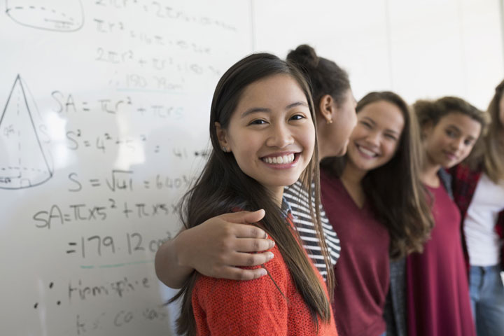 A female student stands sideways, smiling at the camera. She is next to other students, one with an arm around her shoulder. A whiteboard with mathematical work is visible in the background.