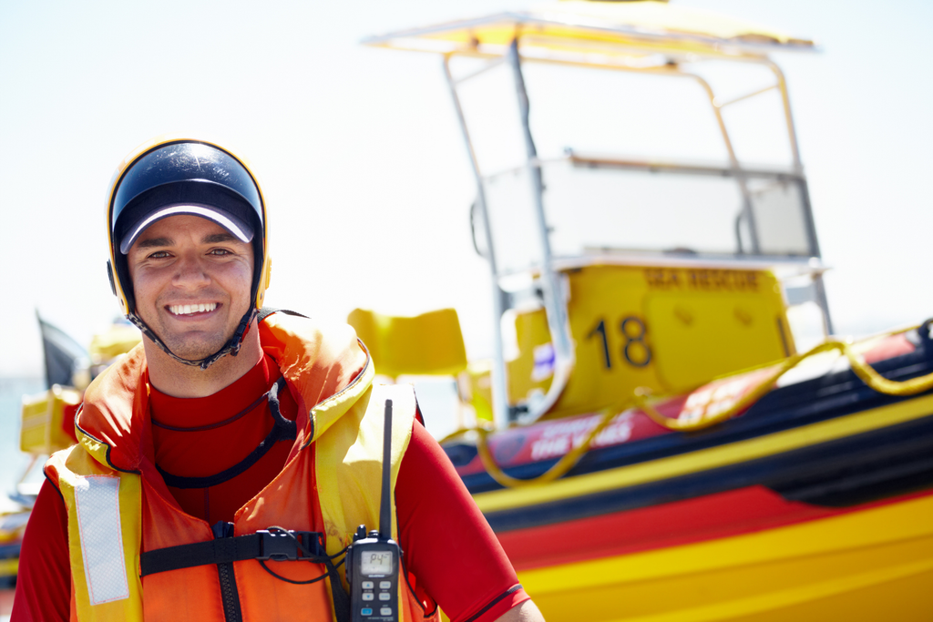 Smiling lifeguard in safety gear standing in front of a rescue boat, ready for duty on a bright, sunny day.