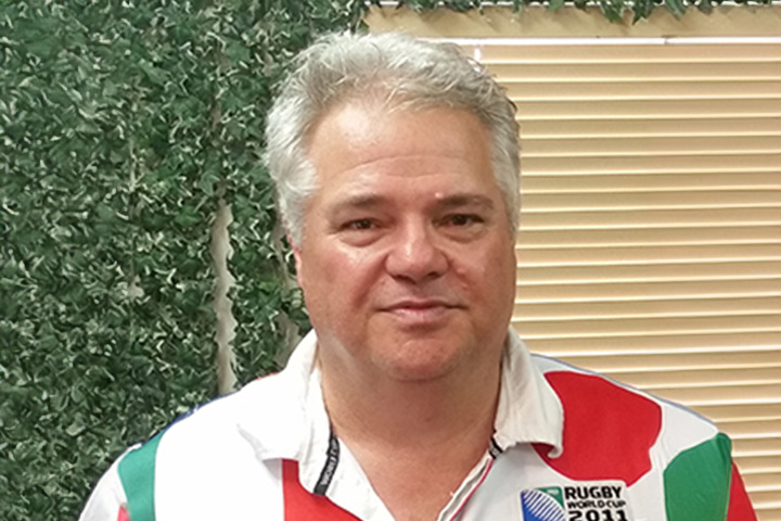 TAFE NSW Engineering teacher Simon Zinader proudly wears a Rugby World Cup 2011 jersey, standing in front of vertical blinds and artificial plants.