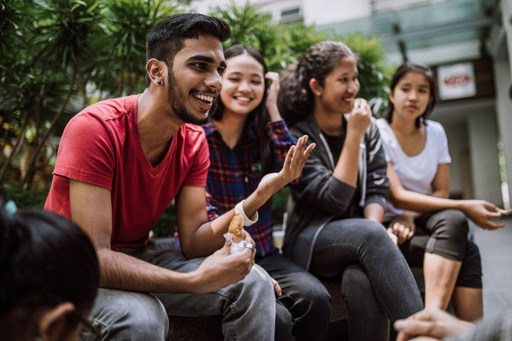A group of students sit together eating lunch and chatting to one another. They’re involved in conversation and the atmosphere is friendly and relaxed.