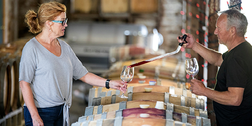 A woman and man stand on either sides of wine barrels. The man reaches across using a glass extraction phial to pour red wine into the win glass held out by the woman.