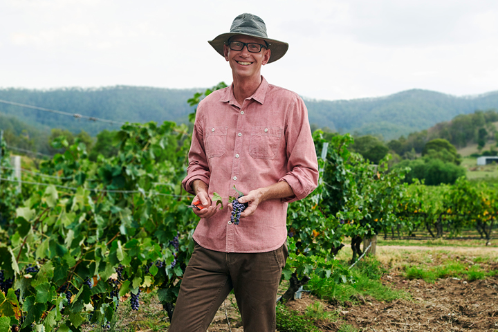 A man stands smiling proudly at the camera, holding a bunch of grapes. He’s surrounded by a lush and healthy vineyard. He’s wearing a bright collared shirt, long, brown pants and boots, and a wide-brimmed sun hat.