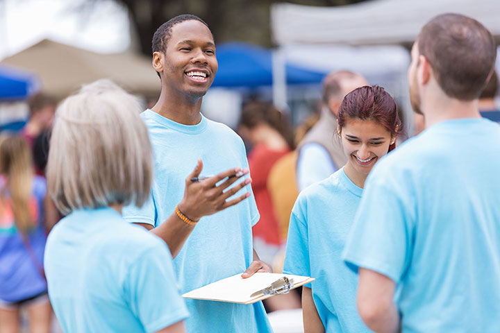 A group of volunteers in light blue shirts are gathered outdoors. One man, holds a clipboard, smiling and speaking to the others. A woman next to him is looking down and smiling. 
