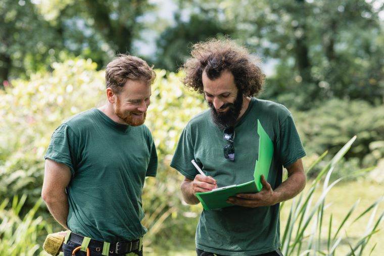 Two men wearing green shirts stand outdoors in a lush, green environment. One man is writing on a green folder while both smile and appear to be engaged in discussion.