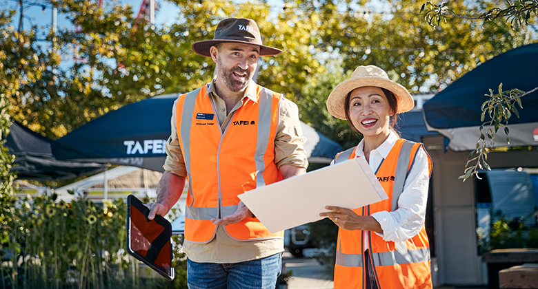 Two people wearing TAFE-branded high-visibility vests and hats, discussing outdoors. One holds a tablet, while the other holds a document folder.