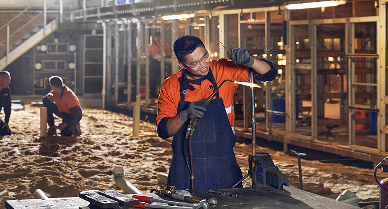 A tradesman working at night with specialised equipment and tools. Other workers are in the background, kneeling and working. The outdoor space has a semi-constructed building lit up with strong outdoor lights.  