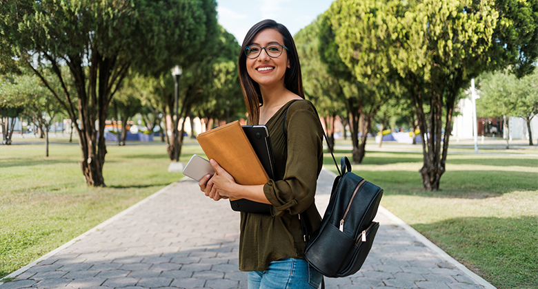 A female student standing in the middle of a pathway, smiling at the camera. She has her work folders in her arms and a backpack over her shoulder. Trees line the path on either side.