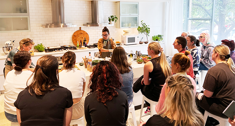A group of women sit in a modern kitchen setting, watching two chefs demonstrate cooking techniques. The room is bright with natural light from large windows, and the chefs are surrounded by fresh herbs and ingredients. The kitchen features sleek white tiles, stainless steel appliances, and wooden chopping boards. The audience observes the chefs closely as they prepare food on a counter.