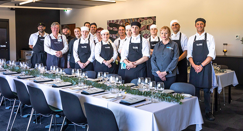 A group of chefs and students from TAFE NSW, dressed in black aprons and chef uniforms, stand proudly behind a beautifully set dining table. The table is adorned with elegant tableware and a eucalyptus leaf arrangement, ready for a formal dining event. The group is posing in a well-lit, modern room, with a background featuring artwork and minimal decor.