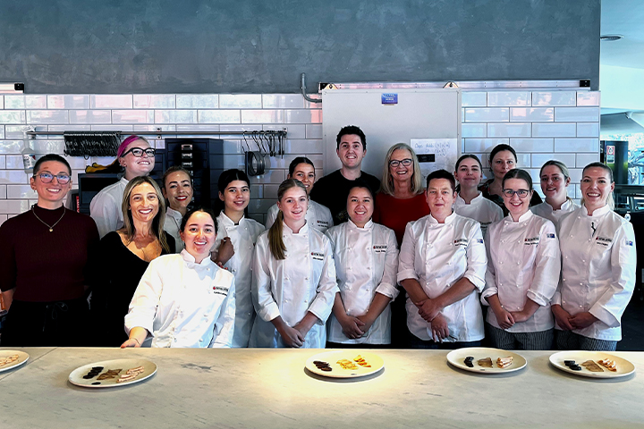 A group of students and teachers pose together in a professional kitchen. The students are dressed in white chef uniforms. There are tasting plates in front of them on a long kitchen bench.