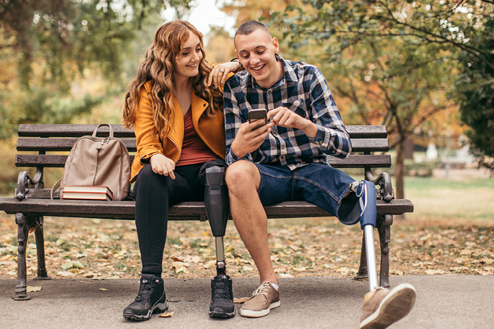 A young woman and young man seated close together, she with one arm resting on his shoulder, both of whom have an artificial left leg, laughing fondly from sharing something on his phone screen.