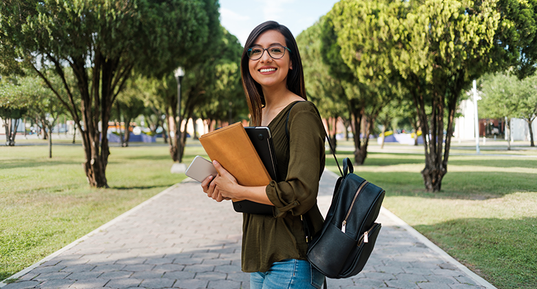 A smiling female student looking out to us from a paved pathway between trees.