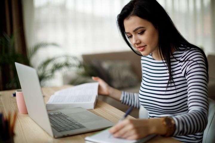 A female student sits at her desk in front of her laptop. She is dressed in a black and white striped top. She is taking notes and has a text book also open on her desk, with a pink coffee mug nearby.