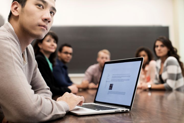 A male student in a beige knitted jumper sits at a table, attentively listening to a presenter. The group, with members from diverse multicultural backgrounds, is actively engaged in the learning environment.