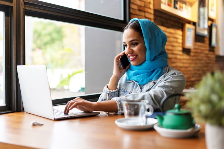 A female student sits at a cafe table with a green teapot and a glass cup nearby. She is wearing a blue hijab and a denim jacket, smiling widely as she takes a phone call and glances at her laptop. A wireless earphone is also visible on the table.