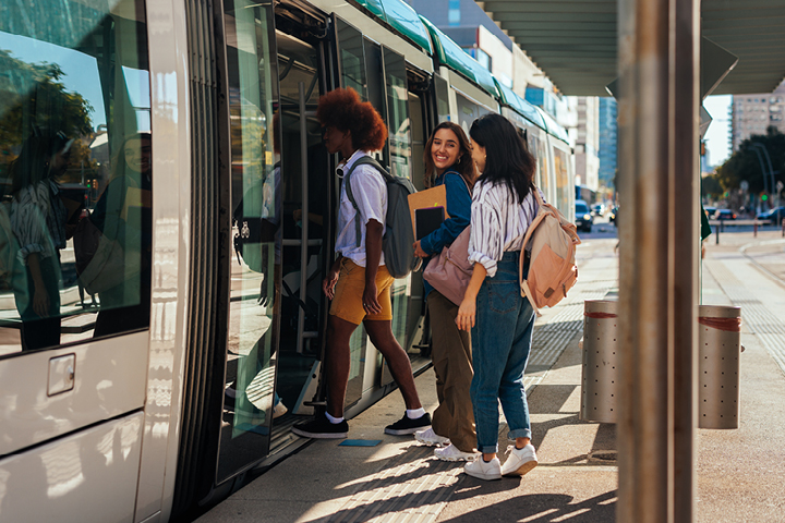 Three happy students carrying books and with backpacks board a train. The sun is out and there are city buildings in the background. They are talking and laughing.