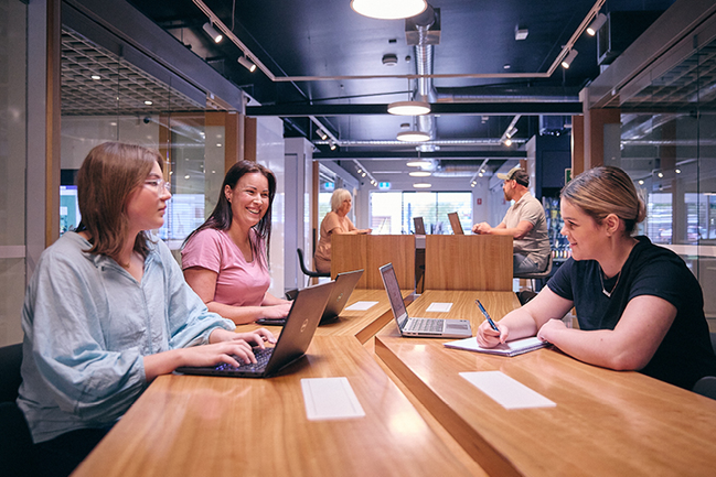 Three smiling people focused on a shared task with open laptop computers. They are in a shared, well-lit connected learning centre space.