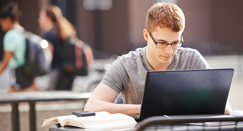 A young man wearing glasses sits in an outdoor area while working on his laptop.