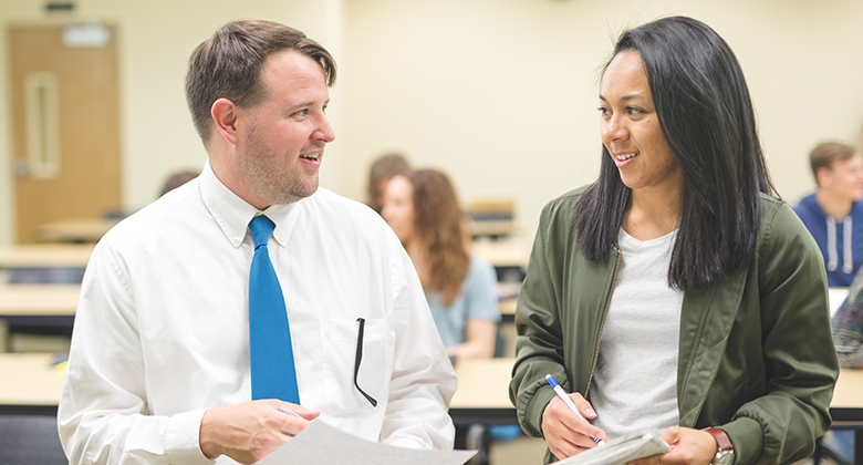 A man and woman engage in conversation in a classroom setting, surrounded by other students and desks.