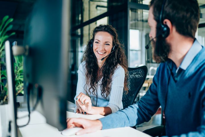 A man and woman are wearing headsets in an office, in front of their computer monitors. They’re sitting at their desks, smiling and having a conversation.