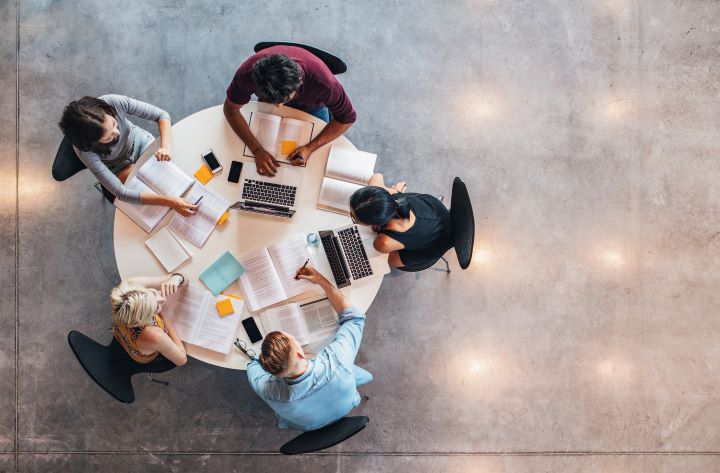 5 university students sitting with their laptops around a circular table. The image has been taken from above and shows papers and books sprawled around the white table.