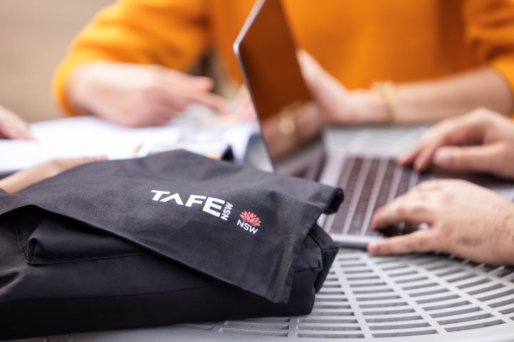 A close-up image showing a TAFE NSW branded black bag on a table. In the background are people working on a laptop and reading documents.