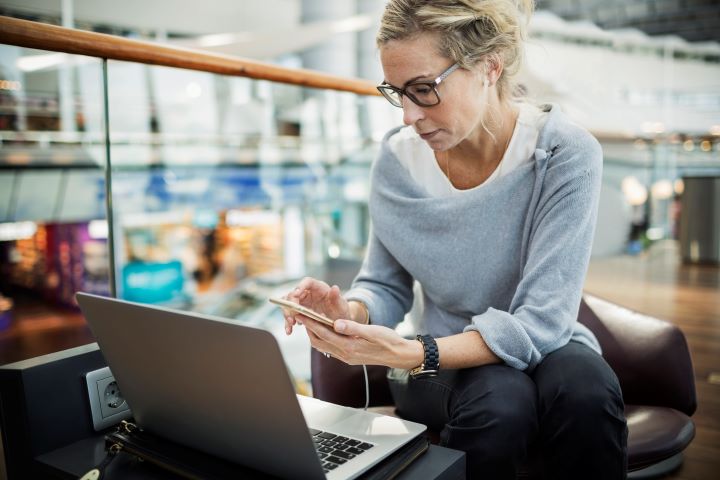 A woman is sitting at her laptop in an office breakout area setting while looking at her phone.