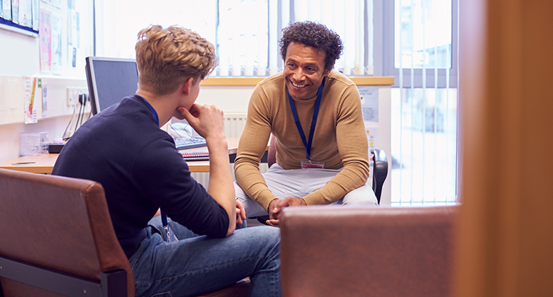 A student sits in an office with a counsellor. The student is facing away from the camera, facing the advisor who has curly hair and wears a lanyard. He is smiling and leaning forward in his chair.
