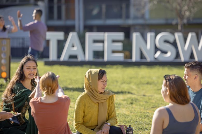 A group of students sit on the grass, talking and relaxing near a large TAFE NSW sign in the background.