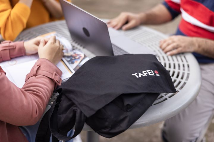 A black TAFE NSW tote bag rests on an outdoor table where two people are engaged in a discussion with a laptop and course materials. 