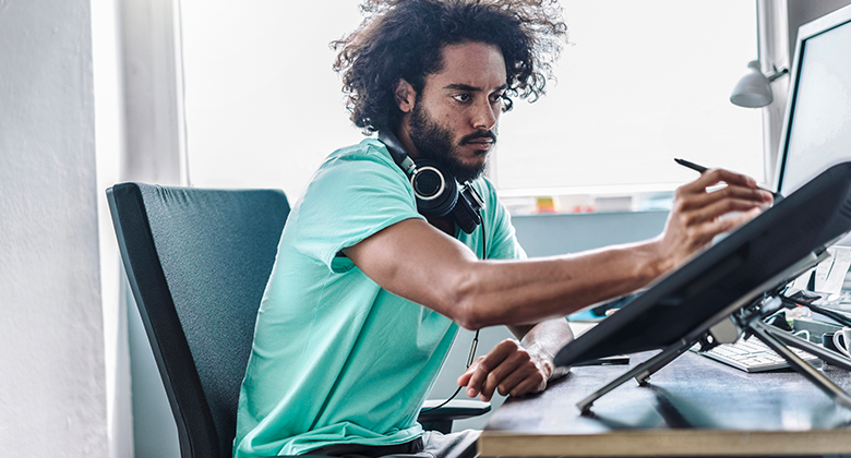 A student at home sits at a desk. He is wearing headphones, holding a stylus and working on a computer tablet.